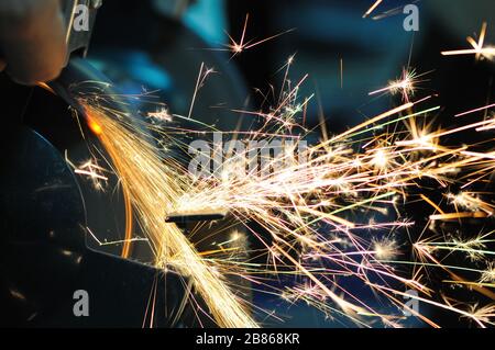 Der Mann hält Details in der Hand und arbeitet mit Geräten zum Schneiden von Metall, helle feurige Funken fliegen auseinander, Nahaufnahme Foto Stockfoto