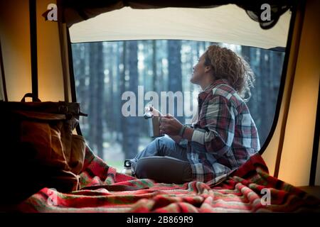 Junge Frau lockig blonde Haare genießen Waldwexkurs beim Trinken eines Tees im Zelt. Freiheit und Abenteuerkonzept Stockfoto