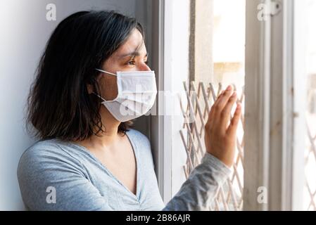 Portrait der jungen Frau, die mit Corona-Virus infiziert ist, mit Gesichtsmaske, die aus dem Fenster schaut, während sie unter Quarantäne steht, mit trauriger und besorgter Ausdrucksweise aufgrund von iso Stockfoto