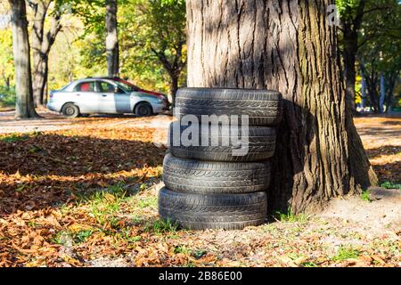 Vier 4 gebrauchte Autoreifen, die neben einem Baum in Parkplätzen gestapelt sind und hinter verschwimmen Autos stehen Stockfoto