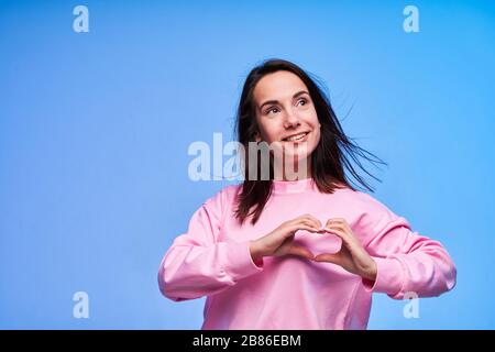 Junge Frau in rosafarbenem Kleid, die Herz mit ihren Händen auf blauem Hintergrund zeigt Stockfoto