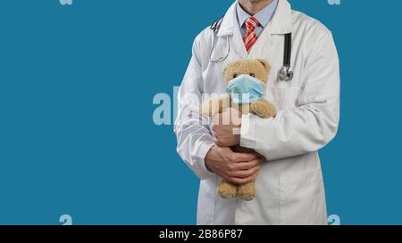 Der Kinderarzt mit einem Stethoskop auf den Schultern hält einen Teddybären in einer medizinischen Maske. Stockfoto