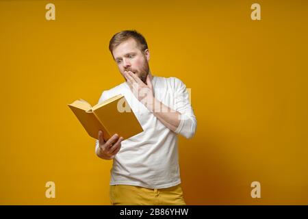 Der junge, bärtige Mann ist fasziniert, ein Buch zu lesen und deckt seinen Mund emotional mit der Hand auf gelbem Hintergrund ab. Stockfoto