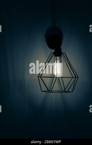 Dunkelblaues Filterfoto der Pendelleuchte mit Lampenschirm in Form von Diamantdraht Karkasse mit Glühbirne innen. Glühfaden in Lampe