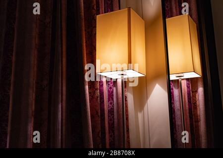 Moderner rechteckiger Lampenschirm und Spiegel an der Wand mit roten verzierten Vorhängen. Goldenes Licht und Dreiecksschatten in der Ecke des Zimmers