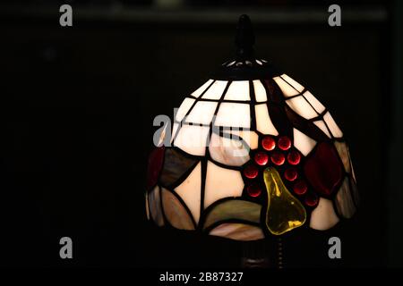 Farbglasleuchter leuchten in Dunkelheit. Tischleuchte auf schwarzem Hintergrund. Mosaik aus weißen und bunten Glasstücken. Stockfoto