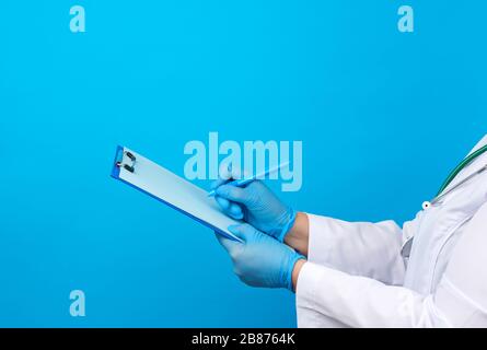 Weibliche Ärztin in einem weißen Mantel halten gummiblaue medizinische Handschuhe einen Ordner für Papiere und schreiben mit der rechten Hand auf ein Blatt, blauer Hintergrund Stockfoto