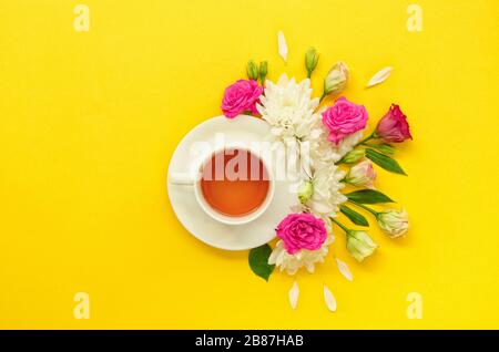 Teezubereitung, eine weiße Tasse Tee, umgeben von rosa gelben, grünen Blumen, die auf gelbem Hintergrund isoliert sind. Horizontales Bild Stockfoto