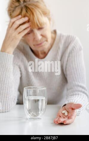 Ältere Frau, die Hand am Kopf hält. Selektiver Fokus auf Pillen und Glas Wasser. Kopfschmerzen- und Stresskonzept Stockfoto
