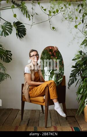 Porträt der schönen jungen Frau in Brillen auf dem Sessel sitzend und posiert bei der Kamera mit Spiegel unter grünen Pflanzen Stockfoto