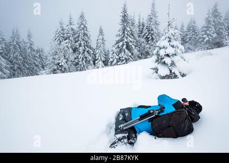 Junger Reisender mit Rucksack fotografiert in einem hohen, schneebedeckten Tannenbaum in hoher Schneeverwehung vor dem Hintergrund von Nebel auf einem frostigen Winter d Stockfoto