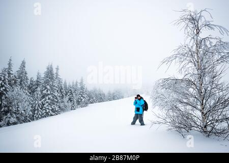 Junger Reisender mit Rucksack fotografiert in einem hohen, schneebedeckten Tannenbaum in hoher Schneeverwehung vor dem Hintergrund von Nebel auf einem frostigen Winter d Stockfoto