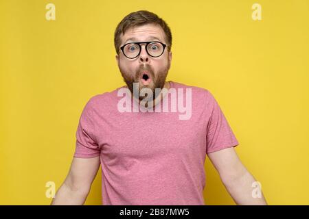 Mann mit Brille im Schock, er öffnete den Mund überraschend, sieht mit großen Augen aus und spreizt die Arme nach den Seiten, auf einem gelben Hintergrund. Stockfoto