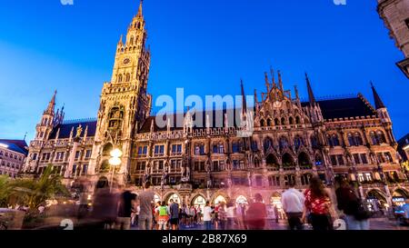 München 2019. Lange Exposition von Touristen, die an einem Sommertag das neue Rathaus bei Sonnenuntergang bewundern und fotografieren. August 2019 in München. Stockfoto