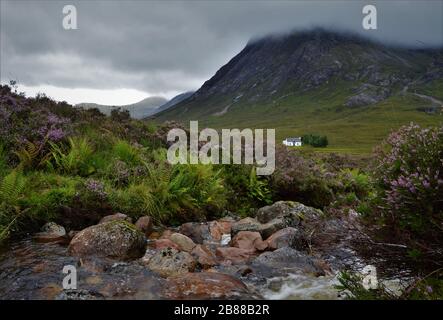 Kleines weißes Haus und kleiner Bach unterhalb von Buachaille Etive Mòr im Glencoe Valley in den schottischen Highlands in Schottland. Stockfoto
