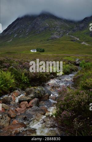 Kleines weißes Haus und kleiner Bach unterhalb von Buachaille Etive Mòr im Glencoe Valley in den schottischen Highlands in Schottland. Stockfoto