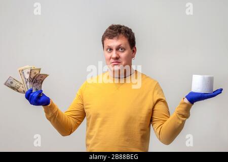 Ein junger Mann in Handschuhen hält Toilettenpapier und eine Packung Dolare und wiegt dabei ihren Wert. Hellgrauer Hintergrund. Europäischer Kerl. Coronavirus und Kovid-19-Konzept Stockfoto