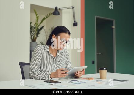 Junge, ernsthafte Geschäftsfrau in Brillen, die an ihrem Arbeitsplatz sitzen und digitales Tablet im Büro verwenden Stockfoto
