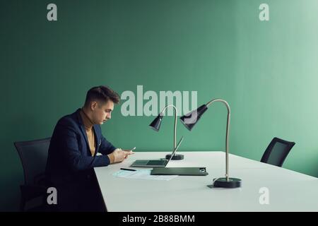 Junger, ernsthafter Geschäftsmann, der an seinem Arbeitsplatz vor dem Laptop sitzt und im Büro mit dem Handy telefoniert Stockfoto