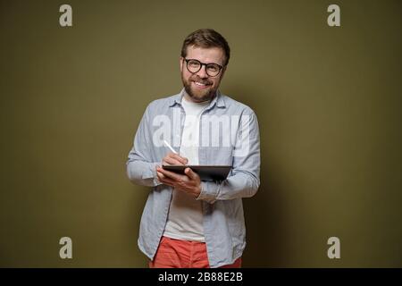 Der freundliche, bärtige Mann in der Brille arbeitet mit einem Tablet- und Stylus Stift, lächelt und blickt auf die Kamera, auf einem grünen Hintergrund. Stockfoto