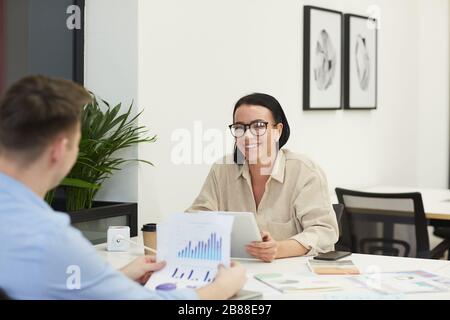 Junge Geschäftsfrau in Brillen lächelt an Geschäftspartner, während sie bei der Besprechung am Tisch über Finanzgrafiken spricht Stockfoto