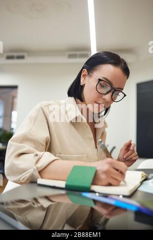 Seriöse junge Geschäftsfrau, die Pläne schreibt und in ihrem Notizblock auflistet, während sie an ihrem Arbeitsplatz sitzt Stockfoto