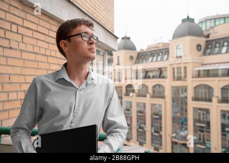 Ein ernsthafter junger Mann in einer Brille, der auf einem Freiluft-Balkon mit Laptop steht und eine Pause hat und nachdenklich aussieht. Flexible Arbeitszeiten und Fernarbeiten Stockfoto