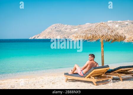Guy in Badehose im Urlaub Mykonos, Mykonos Strand im Sommer mit Sonnenschirm und luxuriösen liegen, blaues Meer mit Berg am Elia Strand Stockfoto