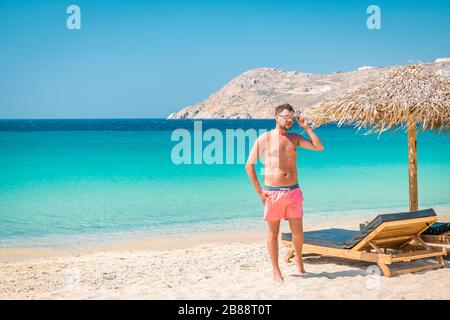 Guy in Badehose im Urlaub Mykonos, Mykonos Strand im Sommer mit Sonnenschirm und luxuriösen liegen, blaues Meer mit Berg am Elia Strand Stockfoto