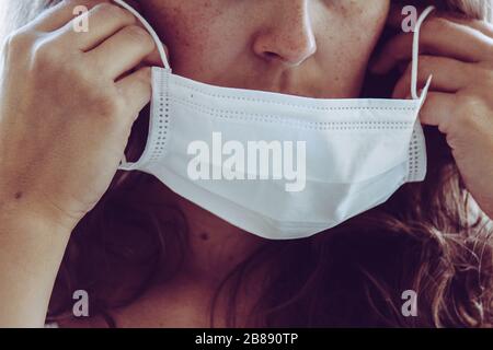 Detail des nicht erkennbaren Gesichts einer Frau, das eine weiße medizinische Gesichtsmaske trägt. Teilweise verschwommen an den Rändern, leichte Körnung. Rotkopfmädchen mit Freckeln. Coronavirus, COVID-19 Quarantäne. Arzt, Pflegekonzept. Stockfoto