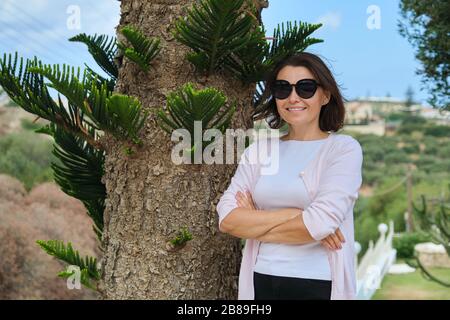 Schöne Frau mittleren Alters mit gekreuzten Armen, Frau in Sonnenbrille in der Nähe von tropischem Baum, Kopierraum. Erfolgreiche Frau, die sich im Resort ausruht Stockfoto