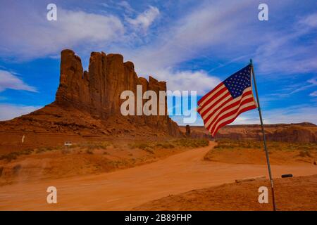 Atemberaubender Blick auf das Kamel butte im Monument Valley. Die amerikanische Flagge wagt am blauen Himmel, erstaunliche rote Felsen und orangefarbene Schmutzstraße, typisch für t Stockfoto