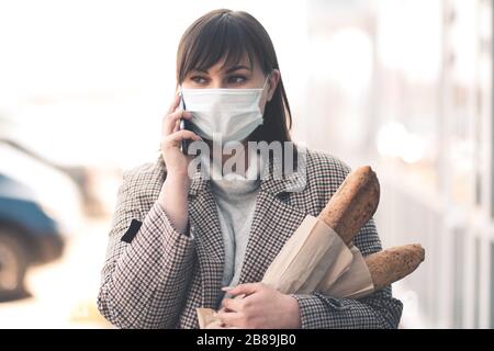 Junge Frau, die medizinische Maske trägt, telefoniert und Lebensmittel auf der Straße im Nahbereich hält. Wegschauen. Virus-Konzept. Soziale Distanzierung. Stockfoto