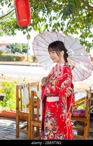 Das Mädchen trägt einen roten traditionellen Kimono, das ist das Nationalkleid Japans und hat einen Regenschirm Stockfoto
