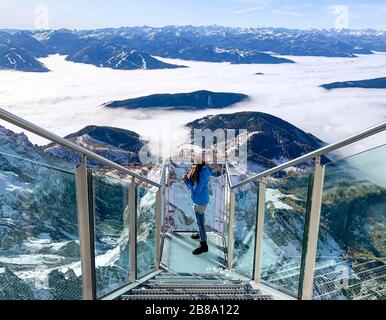 Weibliche Person, die auf einem Skywalk Glass am Dachstein steht und im Winterurlaub den Blick auf die verschneiten Berge in Österreich genießt Stockfoto