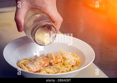 Сlose-up-Koch bestreut große King-Garnelen und Pasta mit Sahnesauce in geriebenem Parmesan aus einem Krug, bei dem Garnelen-Pasta hergestellt werden Stockfoto