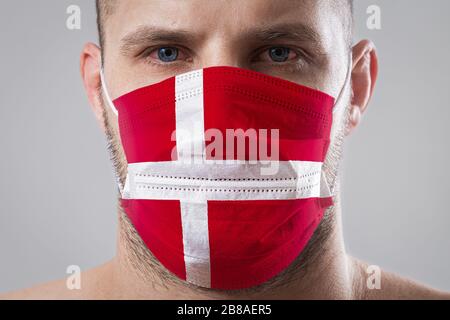 Junger Mann mit schmerzenden Augen in einer medizinischen Maske, die in den Farben der Nationalflaggen von Dänemark gemalt wurde. Medizinischer Schutz vor durch die Luft übertragenen Krankheiten, Korona Stockfoto