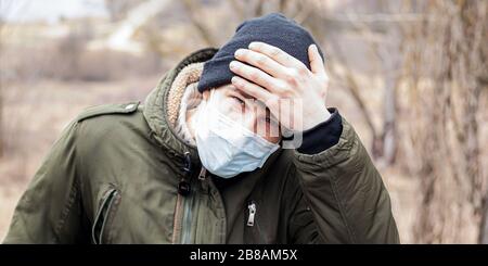 Kerl in einer medizinischen Einwegmaske hält seine Stirn mit der Hand und überprüft die Körpertemperatur. Epidemische Kovid-19 Quarantäne virale Infektion der Atemwege Stockfoto