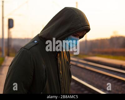 Kerl in einer einmal-medizinischen Maske, die am Bahnhof bei Sonnenuntergang steht. Quarantäne Atemwegserkrankung Epidemie Coronavirus Grippe Public tra Stockfoto