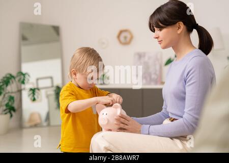 Junge Mutter, die eine Schweinebank hält, während der Sohn eine Münze einlegt, und Mutter, die dem Sohn beibringt, Geld zu sparen Stockfoto