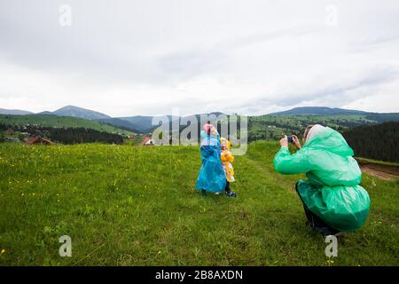 Mama im Regenmantel fotografiert ihre beiden hübschen Kinder in Regenmänteln vor dem Hintergrund malerischer Hügel- und Bergwiesen im Cou Stockfoto