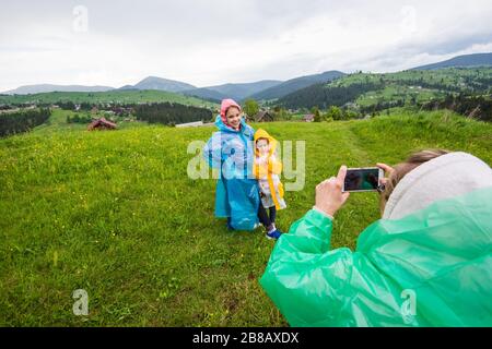 Mama im Regenmantel fotografiert ihre beiden hübschen Kinder in Regenmänteln vor dem Hintergrund malerischer Hügel- und Bergwiesen im Cou Stockfoto