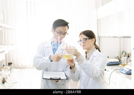Kleines Team von jungen interkulturellen Labormitarbeitern, die über die Eigenschaften von flüssiger Substanz in Röhrchen sprechen, während eines davon Notizen macht Stockfoto
