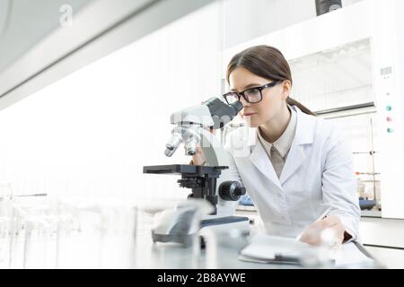 Ernsthafte Forscherin in Whitecoat, die im Mikroskop nach Arbeitsplatz schaut, während sie im wissenschaftlichen Labor Merkmale von Covid19 untersucht Stockfoto