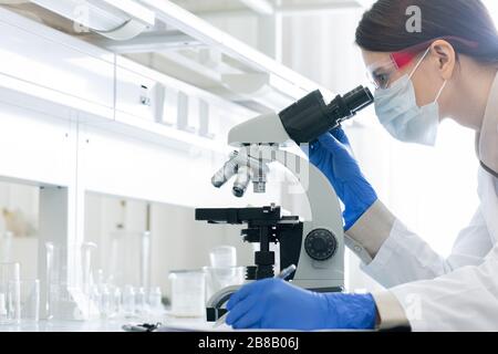 Junge Frau in Whitecoat, Handschuhe, Brille und Maske, die sich über das Mikroskop beugt, während sie im Labor die Eigenschaften von Coronavirus untersucht Stockfoto