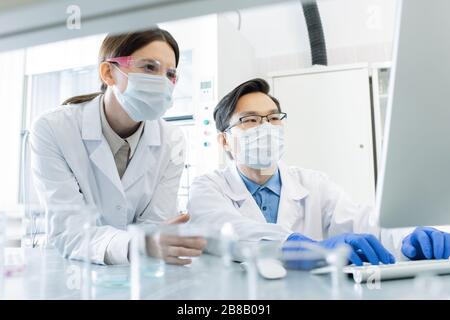 Zwei junge interkulturelle Labormitarbeiter in Masken, Brillen, Handschuhen und Whitecoats, die während der wissenschaftlichen Forschung auf den Computerbildschirm blicken Stockfoto