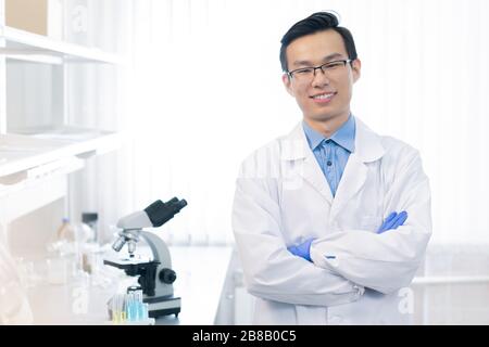 Fröhlicher junger chinesischer Chemiker oder Reseacher mit von der Brust durchzogenen Armen, die am Arbeitsplatz mit Mikroskop vor der Kamera stehen Stockfoto