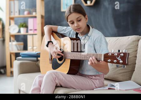 Hübsches Mädchen in der Casualwear auf der Couch sitzend und studierte, um Gitarre zu spielen, während sie Hausunterricht während der Selbstquarantäne nahm Stockfoto