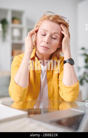 Müde reife Geschäftsfrau in der Casualwear, die den Kopf berührt, während sie am Arbeitsplatz in der häuslichen Umgebung unter Kopfschmerzen leidet Stockfoto