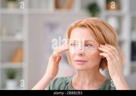 Ziemlich reife Frau mit kurzen blonden Haaren, die Unteraugenflecken setzen und sich dabei um ihr Gesicht während der Selbstisolation zu Hause kümmern Stockfoto